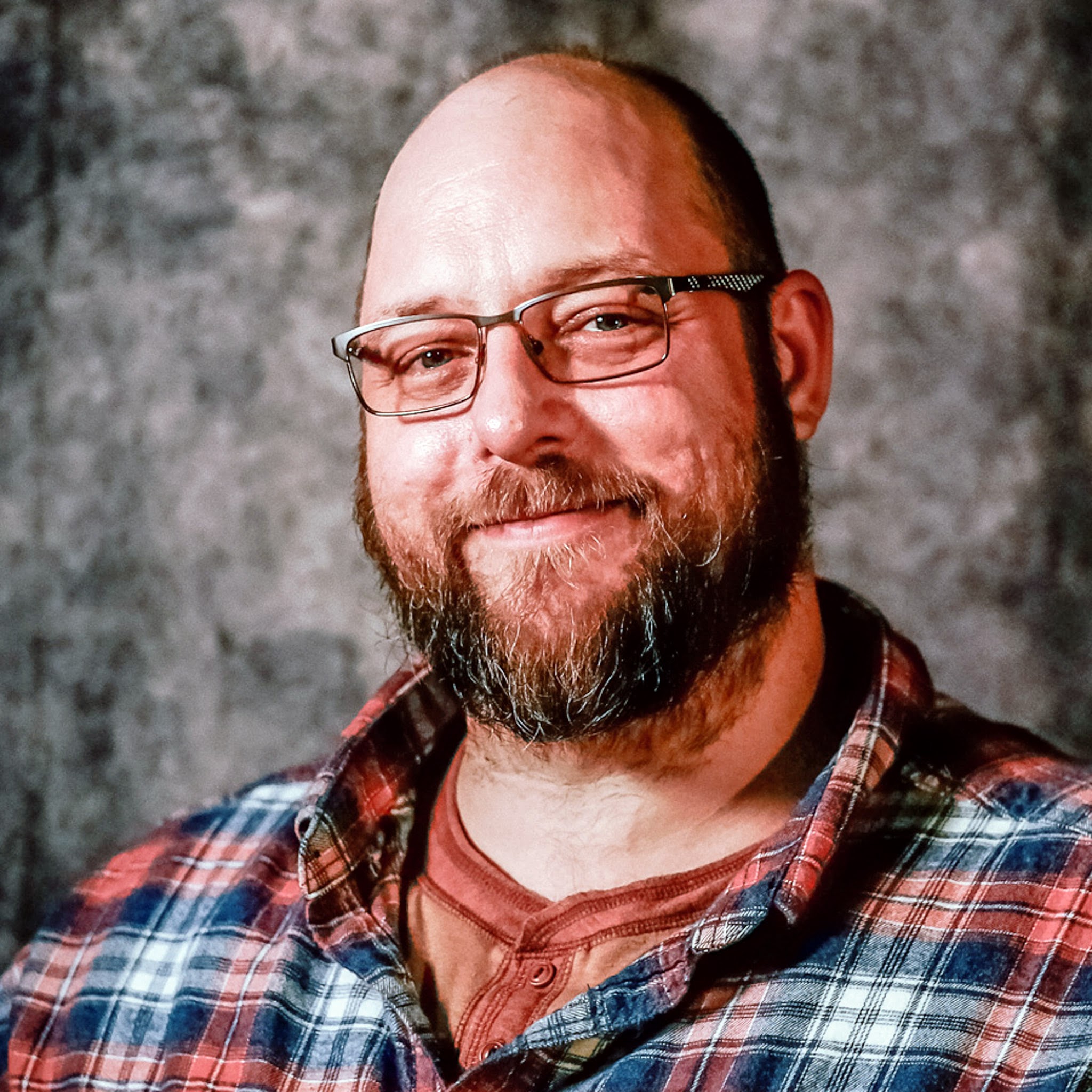 Alvin Kibe posed for portrait photo in red and blue plaid shirt, glasses, with a beard and bald head